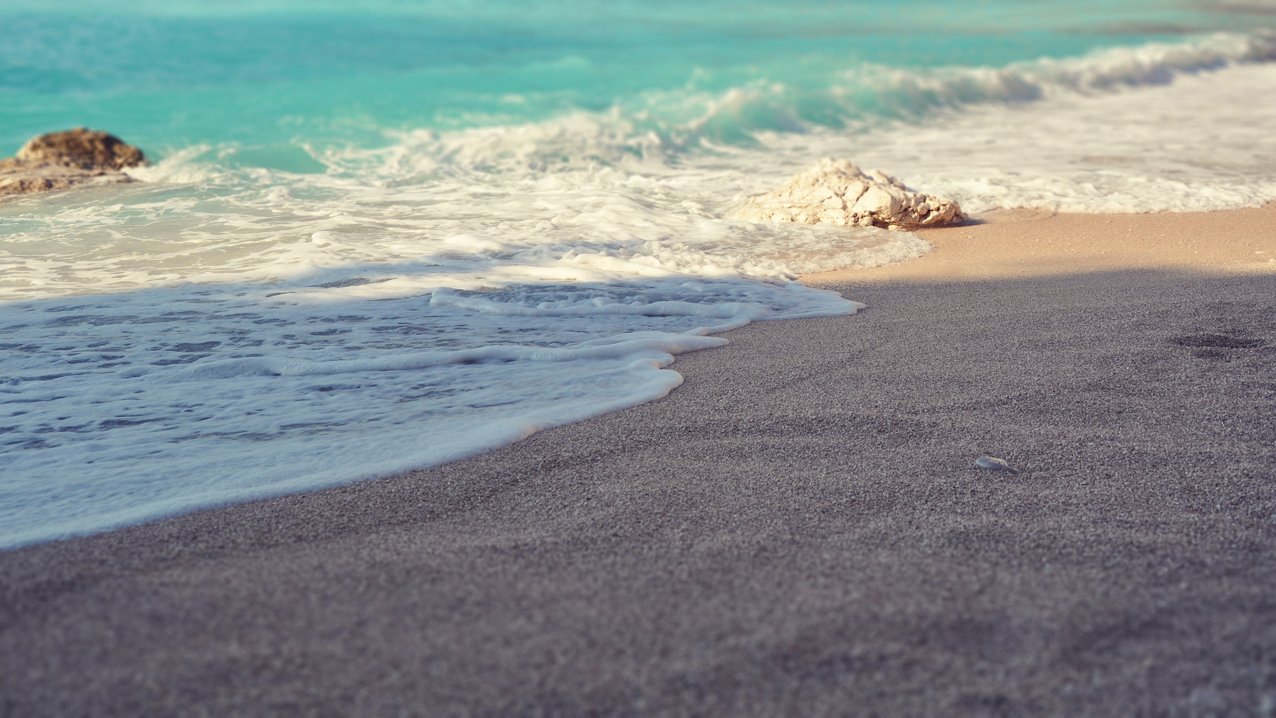 Turquoise waves gently wash the gray sand, large stones lie at the water`s edge, and the sun floods the shore with light.