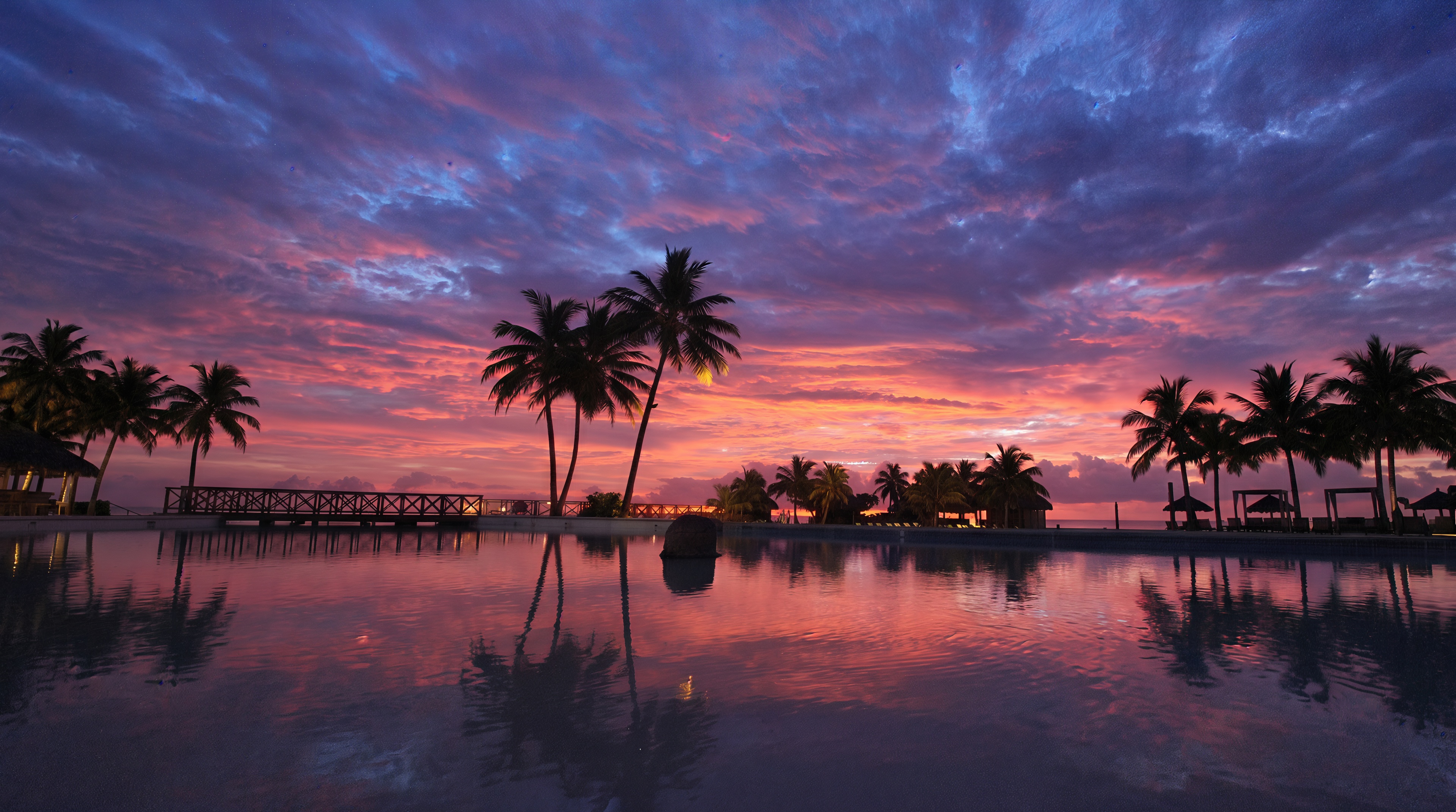 A magnificent tropical sunset in purple and pink tones with silhouettes of palm trees reflected in a mirrored pool.