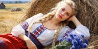 Блондинка с косами в вышитой рубашке лежит на стоге сена, держа букет васильков.