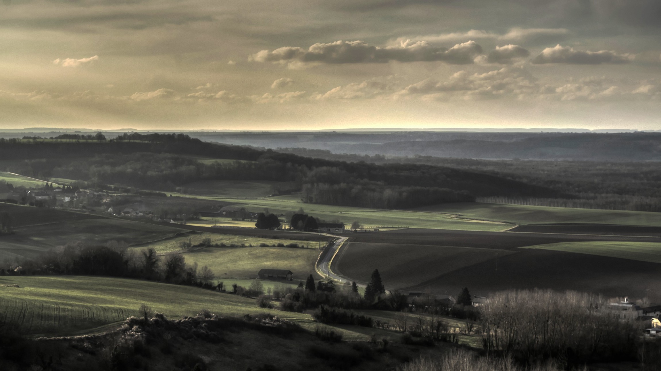 Холмистый пейзаж с полями, извилистой дорогой и облачным небом - умиротворённая сельская панорама.