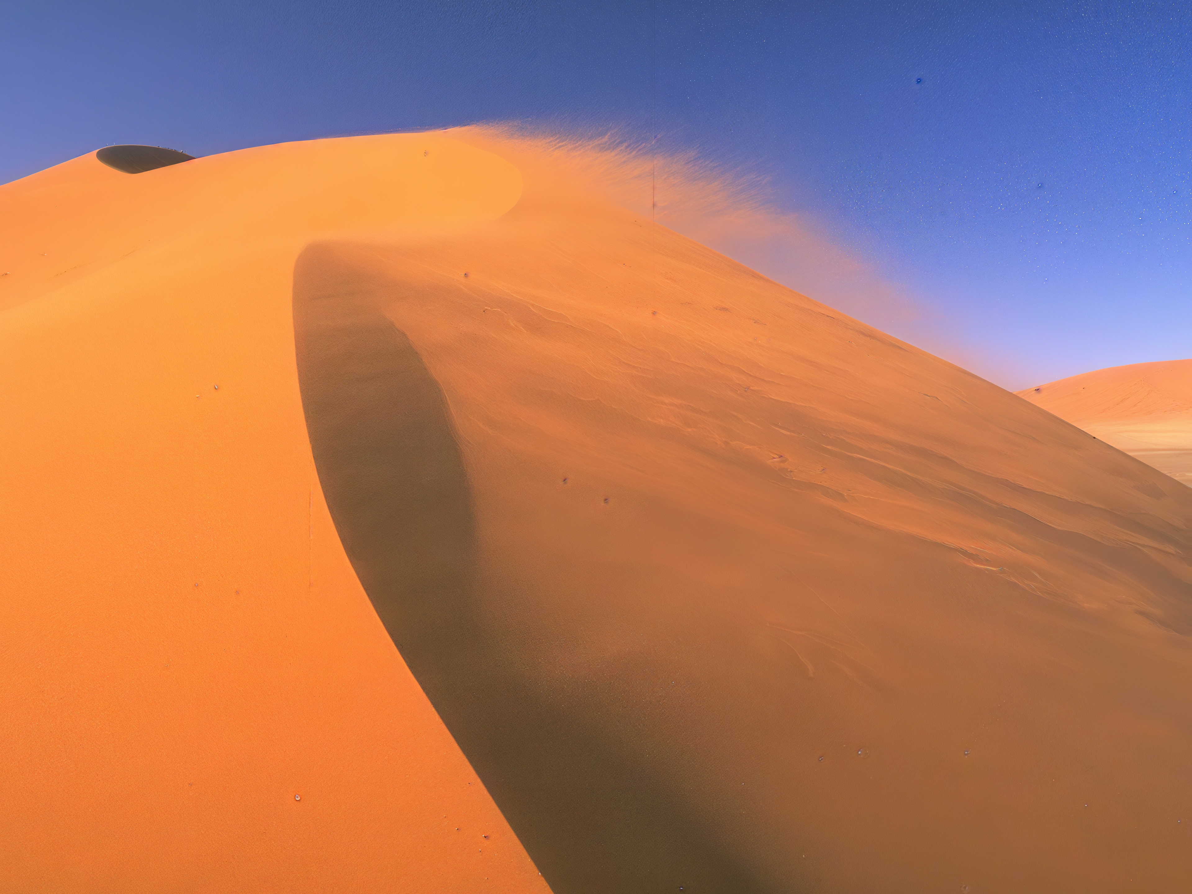 Majestic orange-colored sand dunes under a bright blue sky in the desert, blowing sand in the wind.