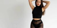 A brunette in a black top and transparent tights poses on a white background.