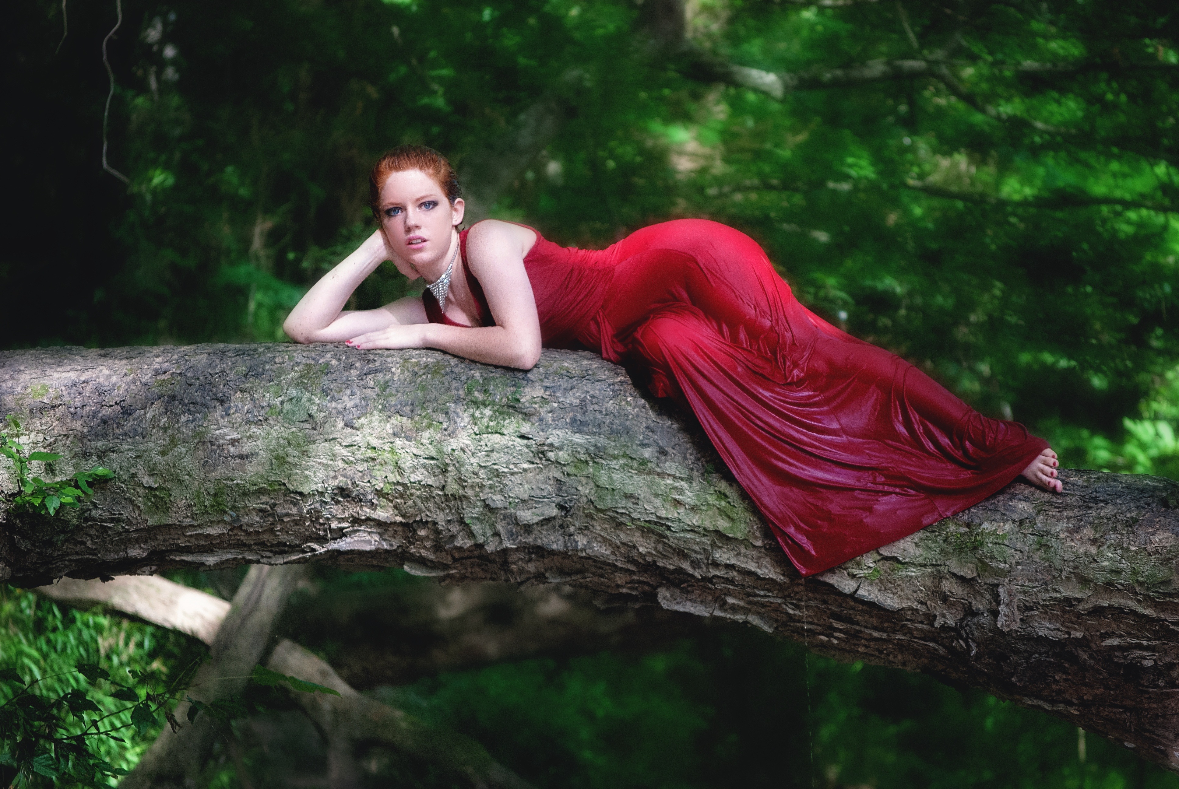Бесплатное фото Рыжеволосая девушка в красном платье лежит на стволе дерева