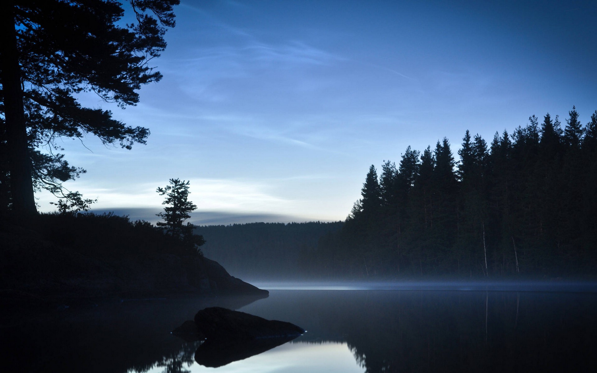 A tranquil forest landscape at dusk with a lake reflecting the blue sky and silhouettes of trees.