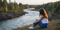 Девушка задумчиво сидит на берегу извилистой реки, окружённой лесом.